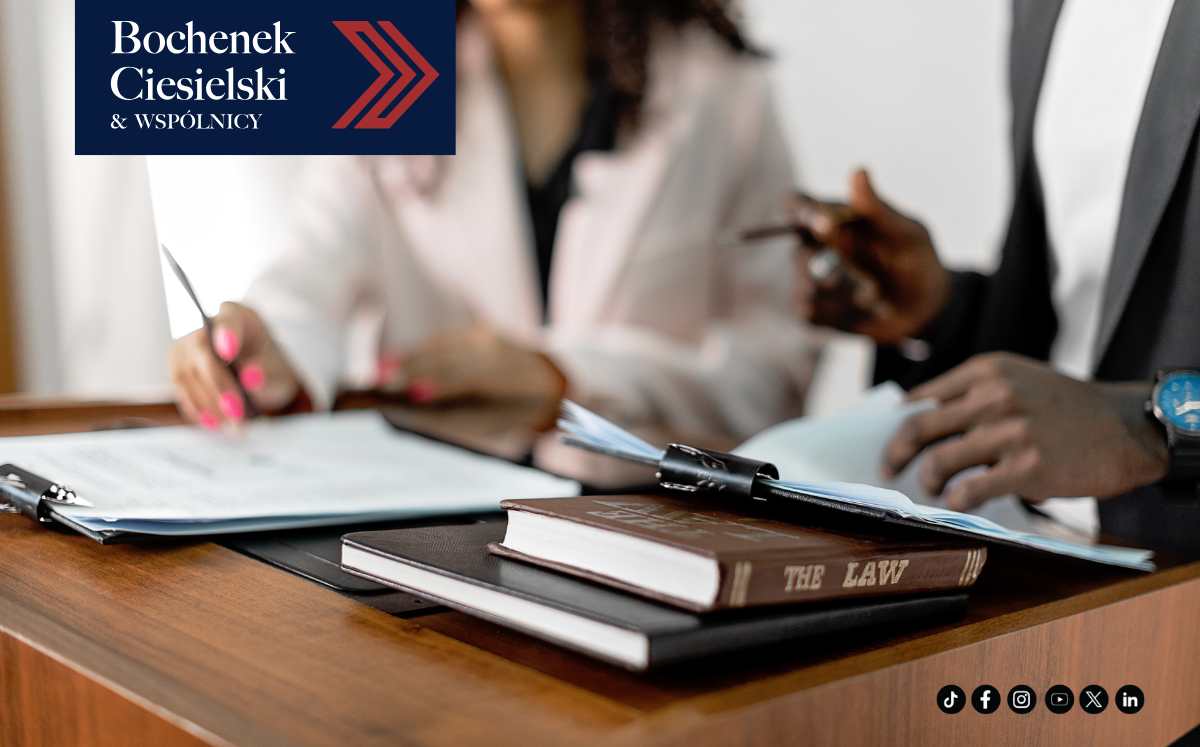 Dwójka prawników omawiających dokumenty przy biurku, na pierwszym planie leżąca książka „The Law”, z logo kancelarii Bochenek Ciesielski & Wspólnicy.