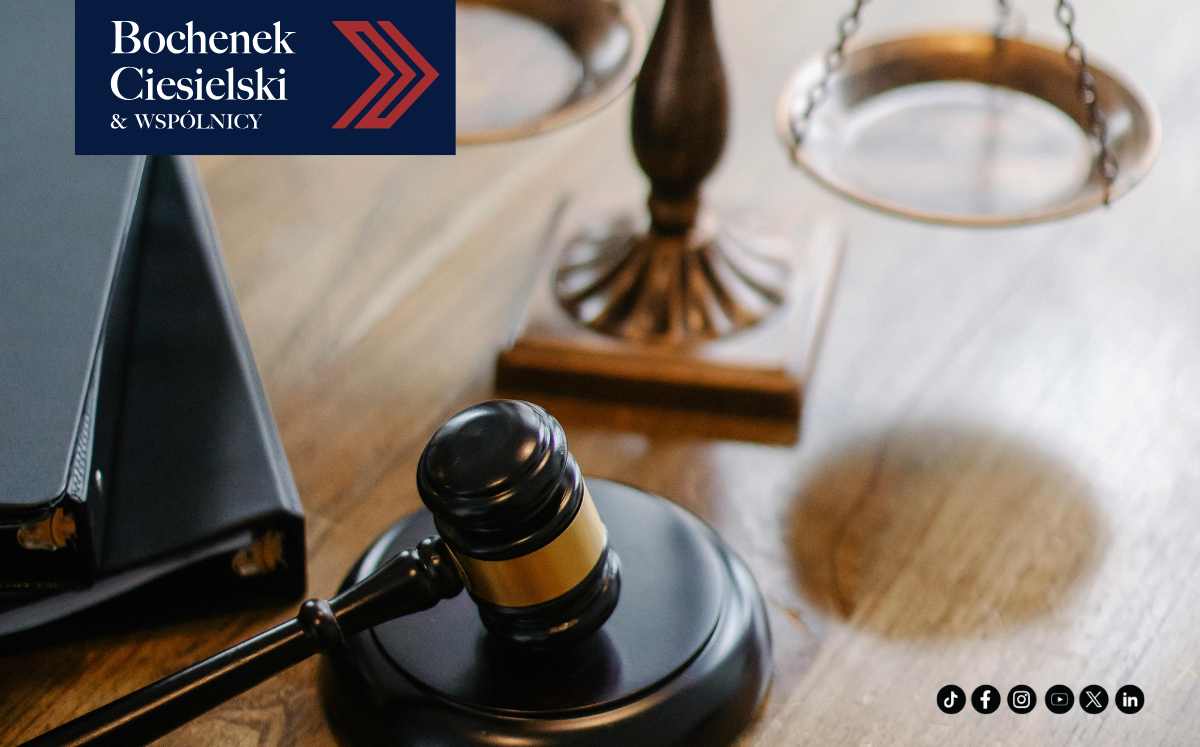 Drewniany stół z młotkiem sędziowskim i wagą sprawiedliwości w tle.