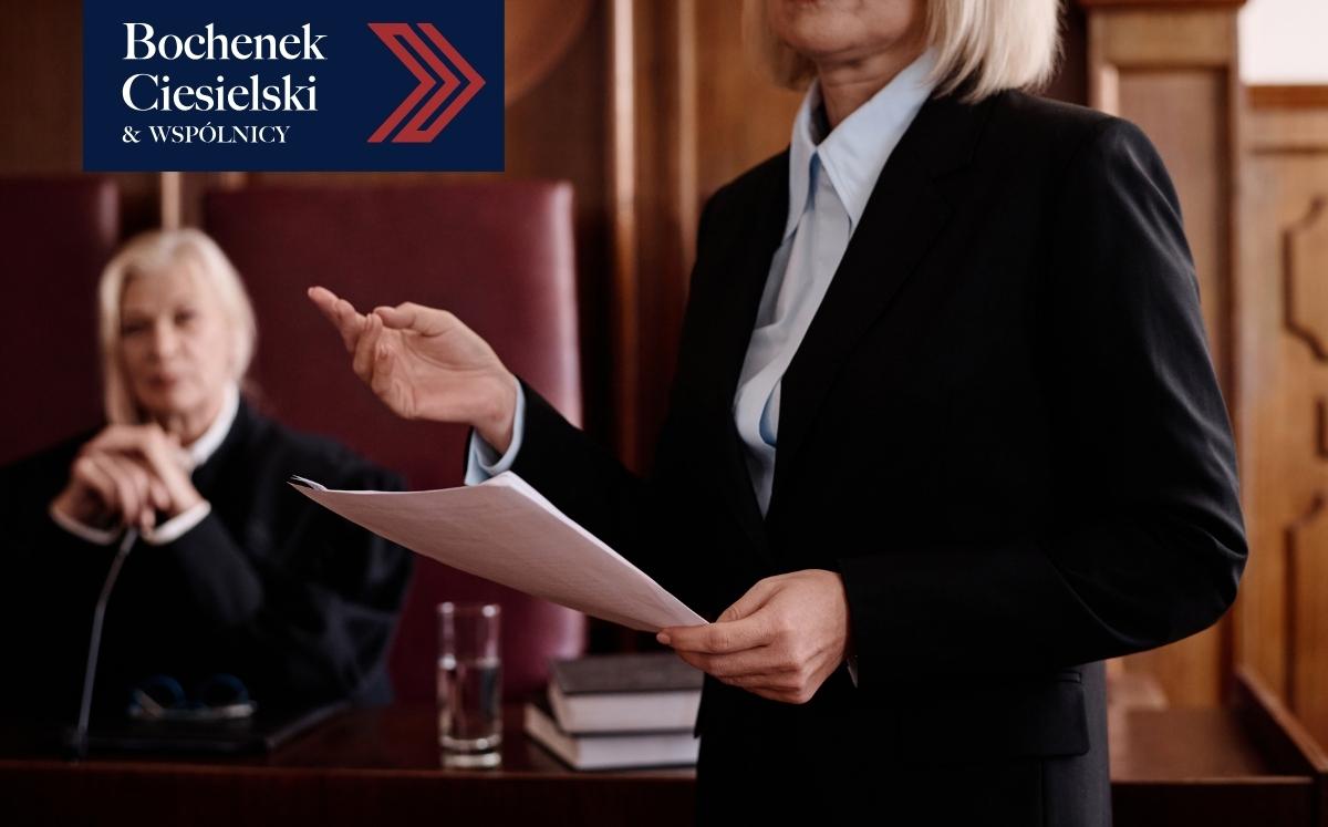 Prawniczka w todze przemawia w sali sądowej, trzymając dokumenty; w tle siedzi sędzia; w lewym górnym rogu logo kancelarii Bochenek Ciesielski & Wspólnicy.