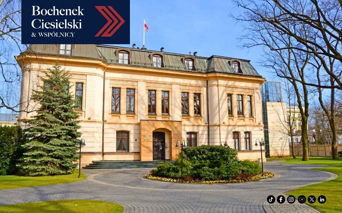Reprezentacyjny, jasny budynek instytucji publicznej otoczony zielenią, sfotografowany w słoneczny dzień; w lewym górnym rogu logo kancelarii Bochenek Ciesielski & Wspólnicy.