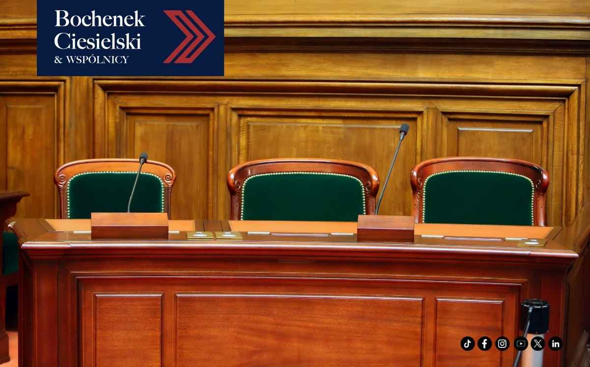 Drewniany stół sędziowski z trzema zielonymi fotelami i mikrofonami w sali sądowej, symbolizujący postępowanie sądowe.