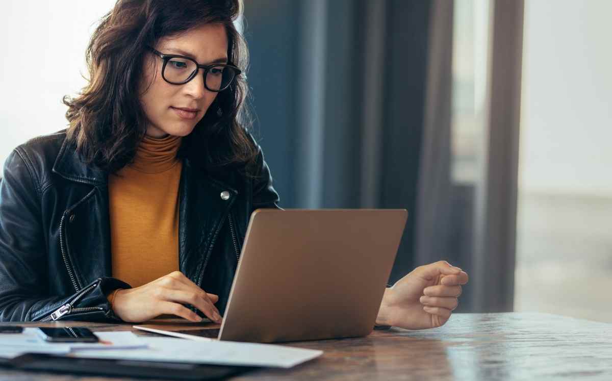 Kobieta w okularach pracuje przy laptopie, siedząc przy drewnianym stole z dokumentami.