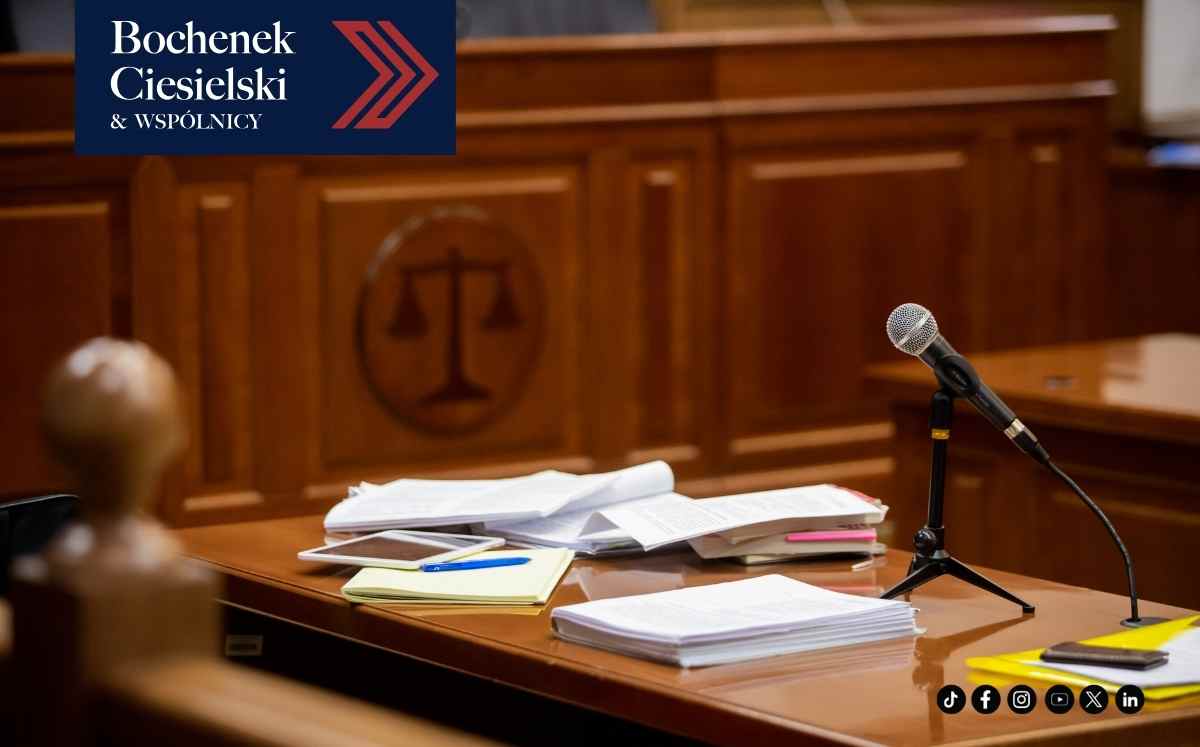 Stół na sali sądowej z dokumentami i mikrofonem na pierwszym planie, w tle drewniana ława sędziowska z symbolem wagi sprawiedliwości oraz logo kancelarii Bochenek, Ciesielski i Wspólnicy.