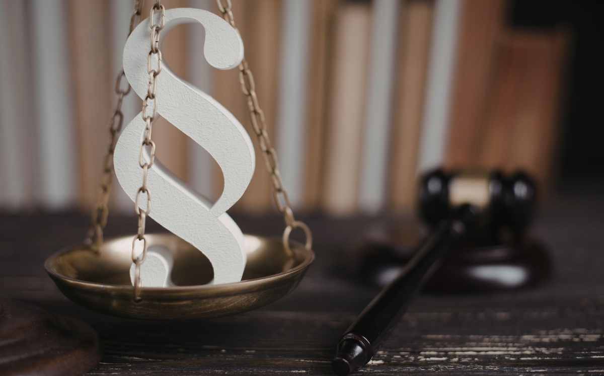 Symbol paragrafu stojący na szali wagi sądowej, w tle młotek sędziowski i regał z książkami prawniczymi.
