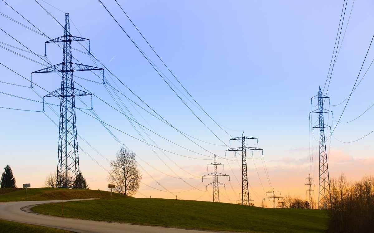 Słupy wysokiego napięcia i linie energetyczne biegnące nad zielonym pagórkiem o zachodzie słońca.
