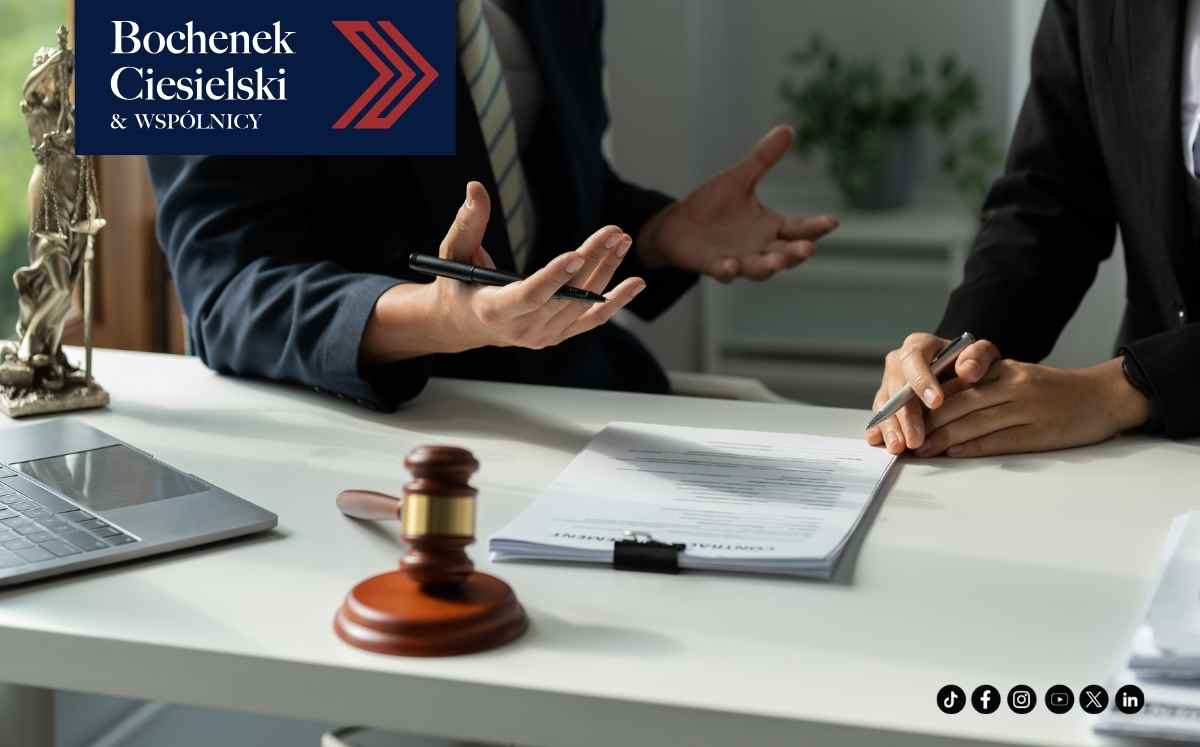 Dwóch prawników omawia dokumenty przy biurku, na pierwszym planie widoczny młotek sędziowski i laptop.