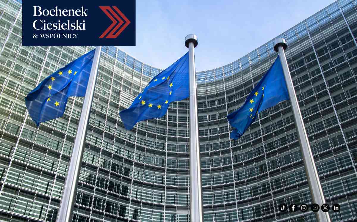 Trzy flagi Unii Europejskiej powiewające na wysokich masztach przed przeszklonym, półkolistym budynkiem instytucji unijnej w Brukseli.