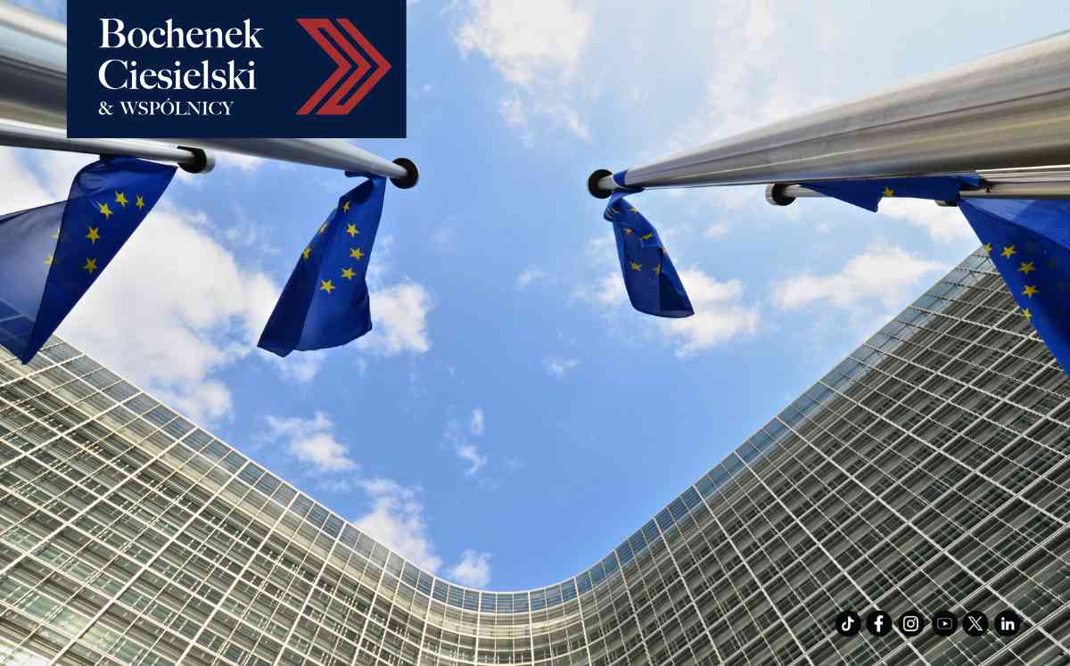 Widok z dołu na maszty z flagami Unii Europejskiej powiewającymi na tle błękitnego nieba z białymi chmurami. Po prawej stronie widoczna przeszklona fasada nowoczesnego budynku.