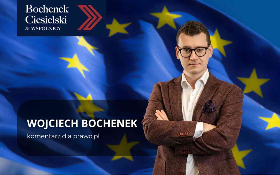 Portret Wojciecha Bochenka w okularach i brązowej marynarce, z założonymi rękami, na tle powiewającej flagi Unii Europejskiej.
