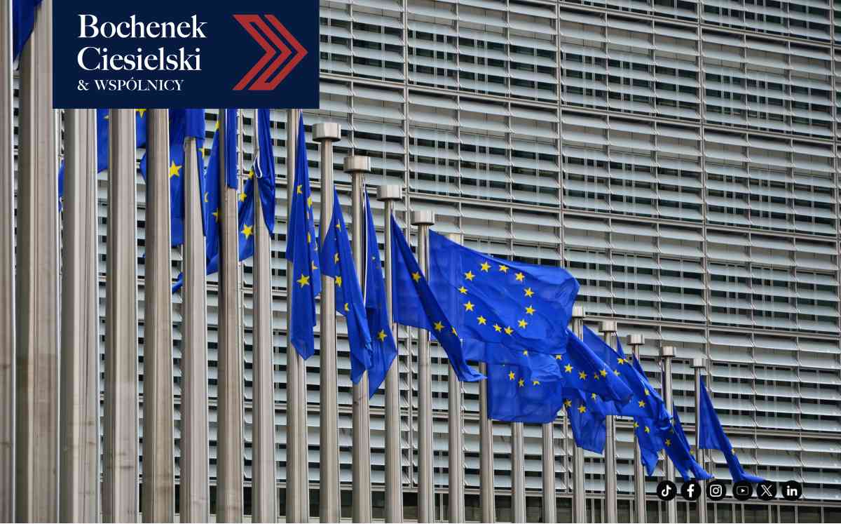 Długi szereg masztów z powiewającymi flagami Unii Europejskiej na tle nowoczesnej, lamelowej elewacji budynku instytucji unijnej.