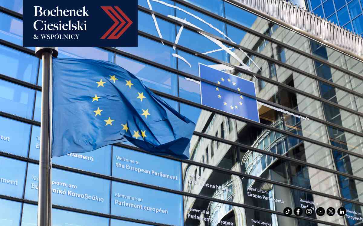 Flaga Unii Europejskiej powiewająca na tle szklanej elewacji budynku Parlamentu Europejskiego, na której widoczne są napisy „Welcome to the European Parliament” w różnych językach.