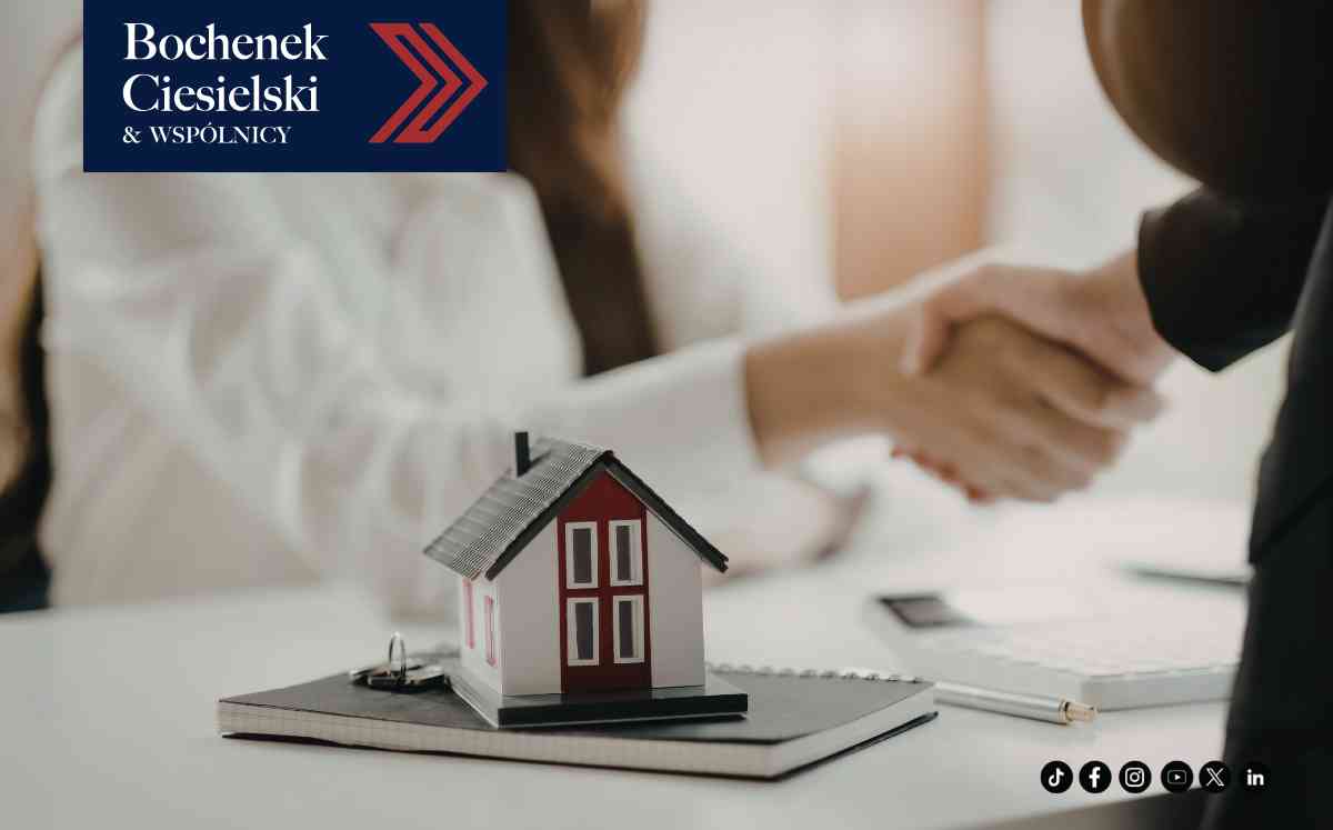 Uścisk dłoni dwóch osób nad biurkiem, na którym stoi makieta białego domu z czerwonym dachem.