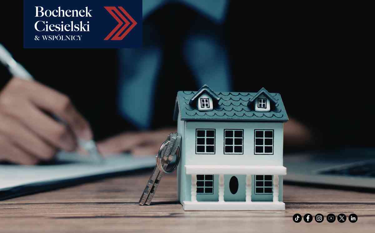 Makieta piętrowego, błękitnego domu z kluczami w zamku, na pierwszym planie, z rozmazanym wizerunkiem osoby piszącej na dokumentach.