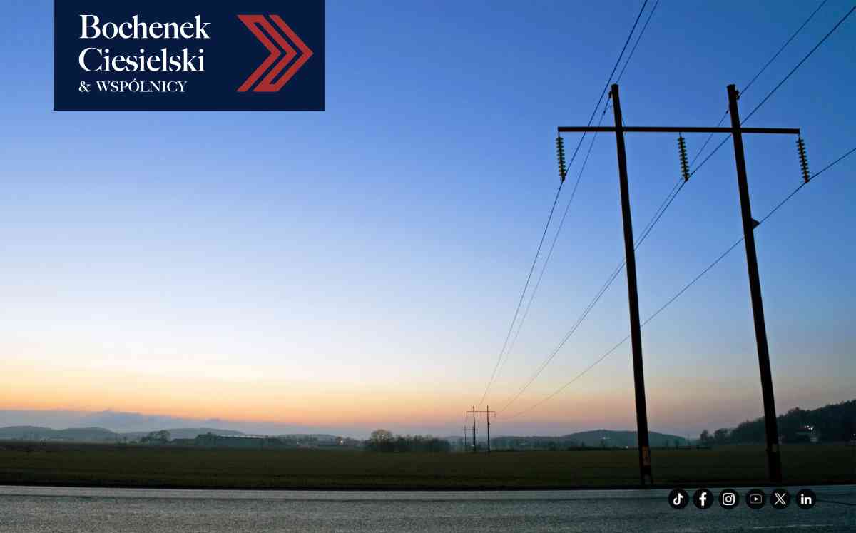 Szereg słupów elektroenergetycznych o różnych konstrukcjach biegnący przez rozległe pole na tle zachodzącego słońca.
