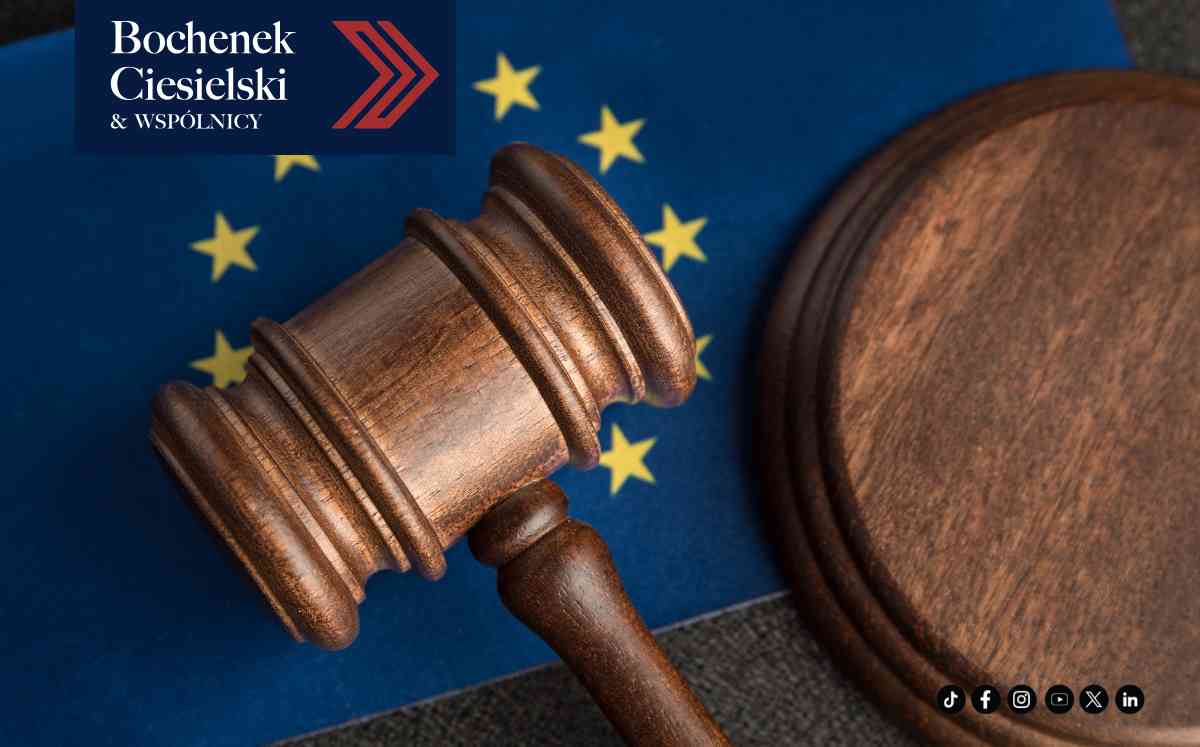 Zbliżenie na drewniany młotek sędziowski leżący obok podstawki na tle flagi Unii Europejskiej z żółtymi gwiazdami.