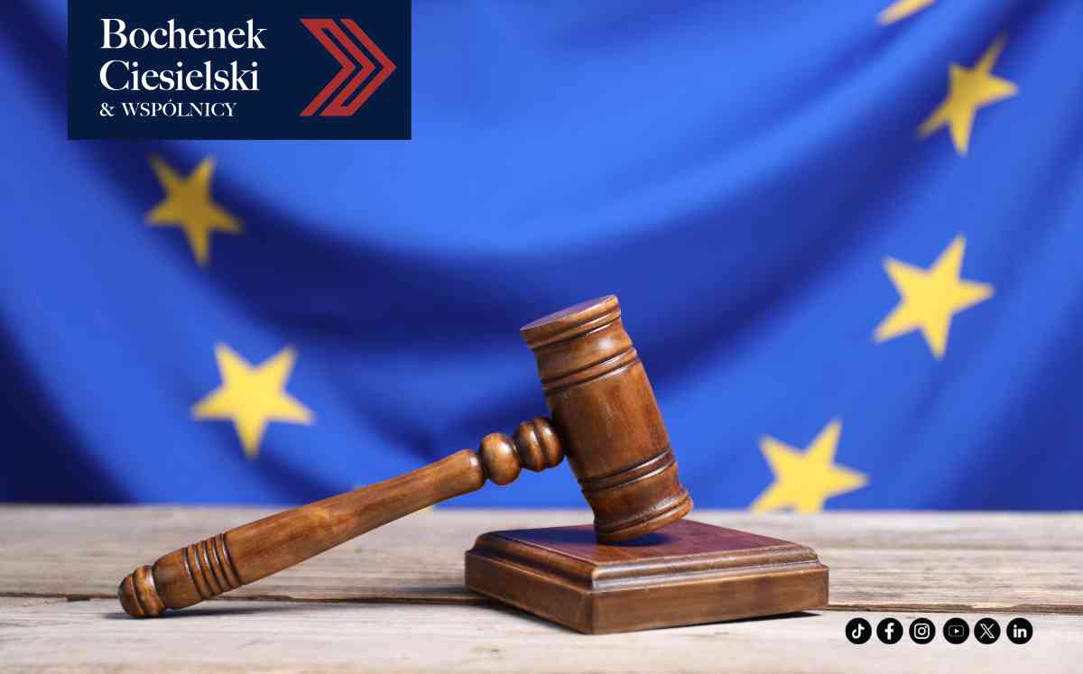 Drewniany młotek sędziowski na podstawce, ustawiony centralnie na jasnym, drewnianym blacie. W tle niebieska flaga Unii Europejskiej z żółtymi gwiazdami.