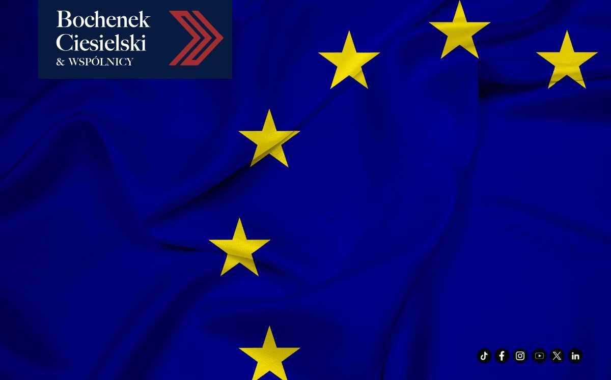 Zbliżenie na powiewającą flagę Unii Europejskiej z żółtymi gwiazdami na niebieskim tle. W lewym górnym rogu znajduje się logo kancelarii.