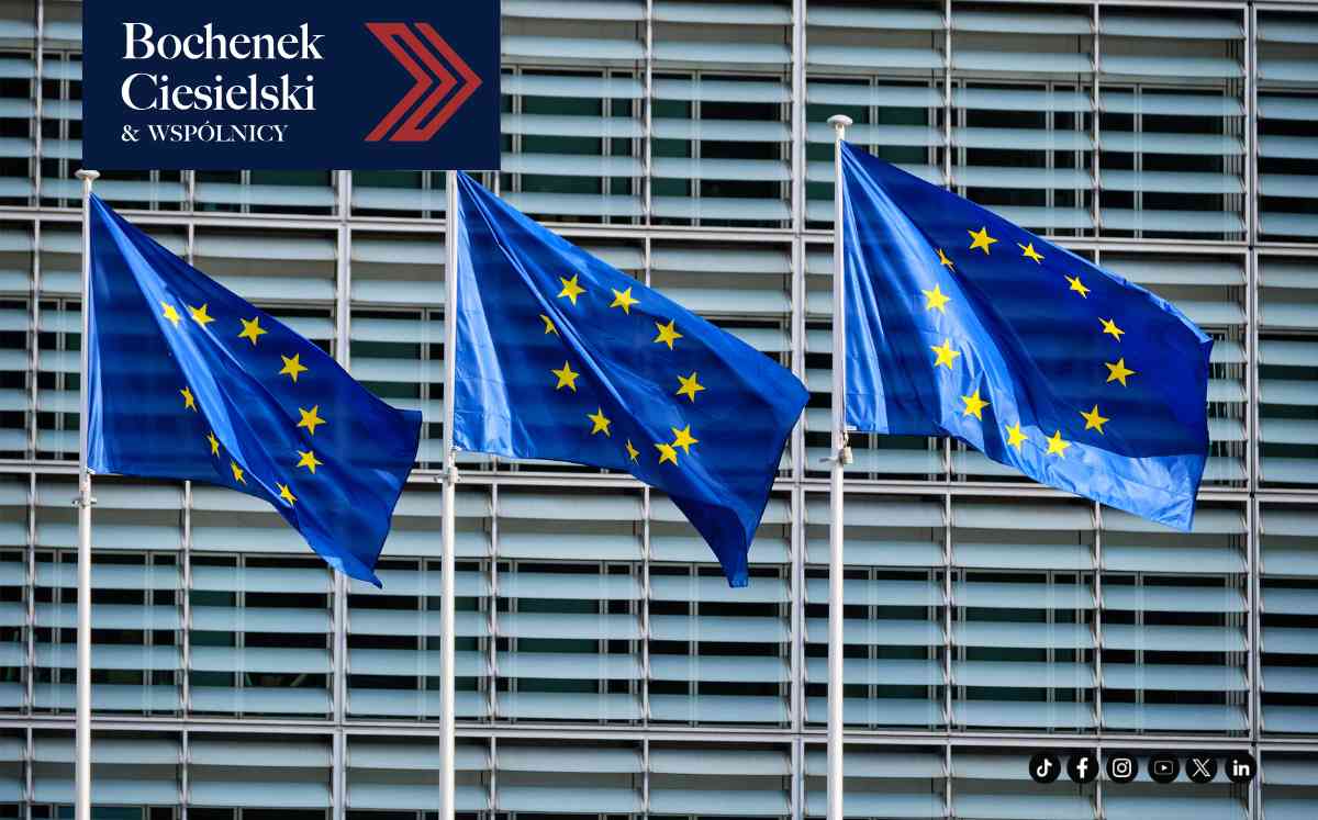 Zbliżenie na drewniany młotek sędziowski leżący obok podstawki na tle flagi Unii Europejskiej z żółtymi gwiazdami.