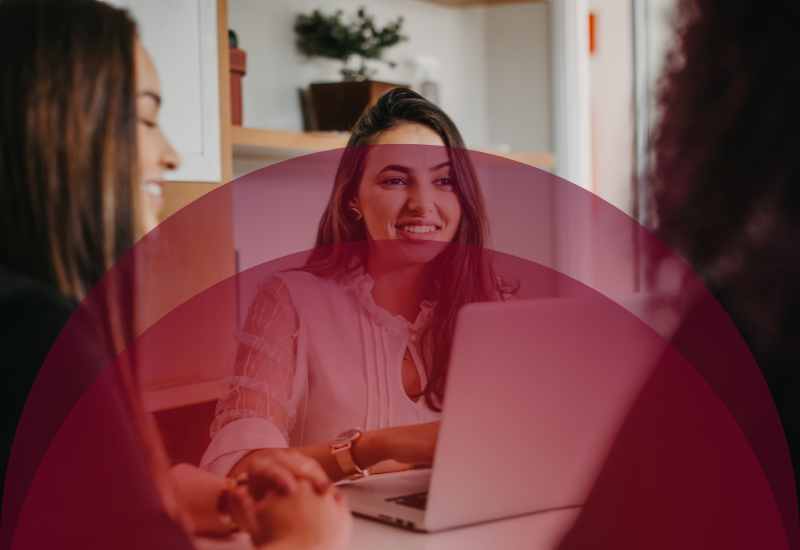 Woman smiles while working on a laptop during a casual group meeting with teammates nearby