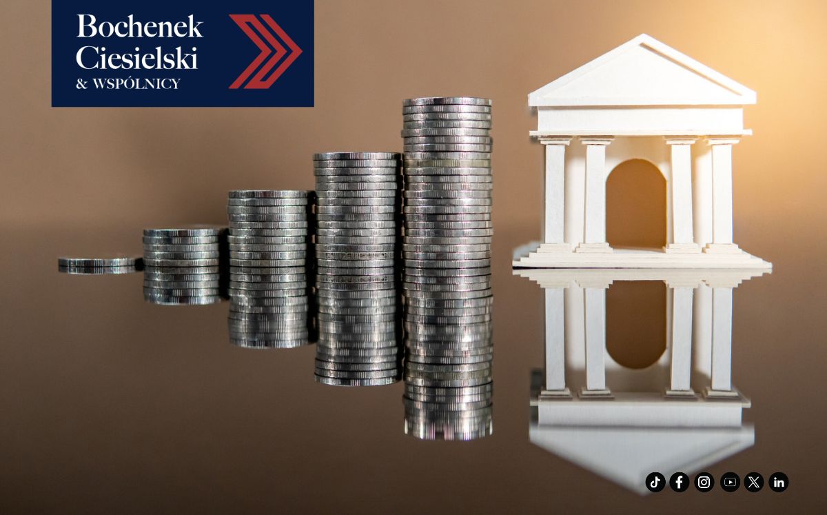 Zbliżenie na figurkę budynku przypominającą bank, obok stoją rosnące w jej stronę kolumny z monet.