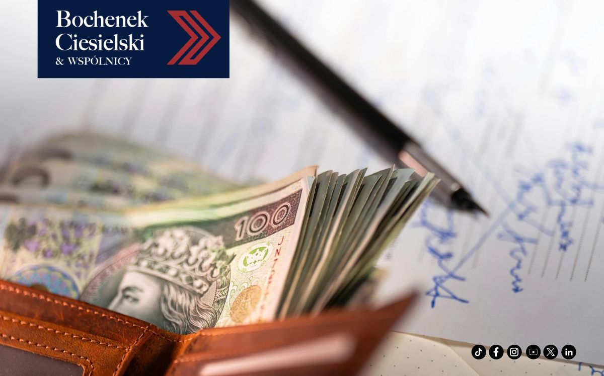 Zbliżenie na otwarty, skórzany portfel, z którego wysuwają się polskie banknoty stuzłotowe. W tle widać podpisany dokument, na którym leży pióro.